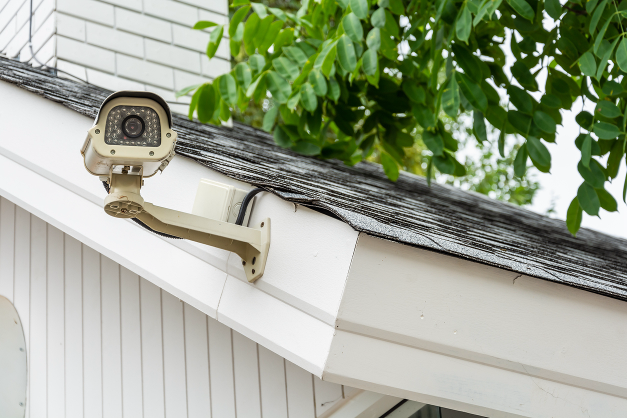 A home security camera mounted on the side of a slanted roof with foliage in the background.